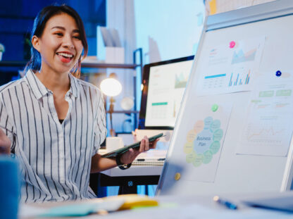 Asia businesswoman social distancing in new normal for virus prevention looking at camera presentation to colleague about plan in video call while work in office night. Lifestyle after coronavirus.