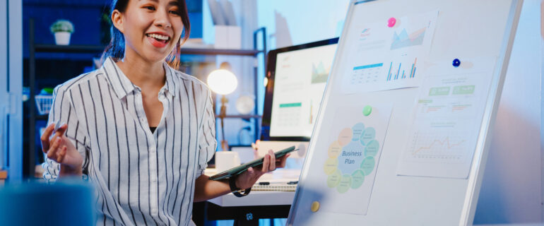 Asia businesswoman social distancing in new normal for virus prevention looking at camera presentation to colleague about plan in video call while work in office night. Lifestyle after coronavirus.